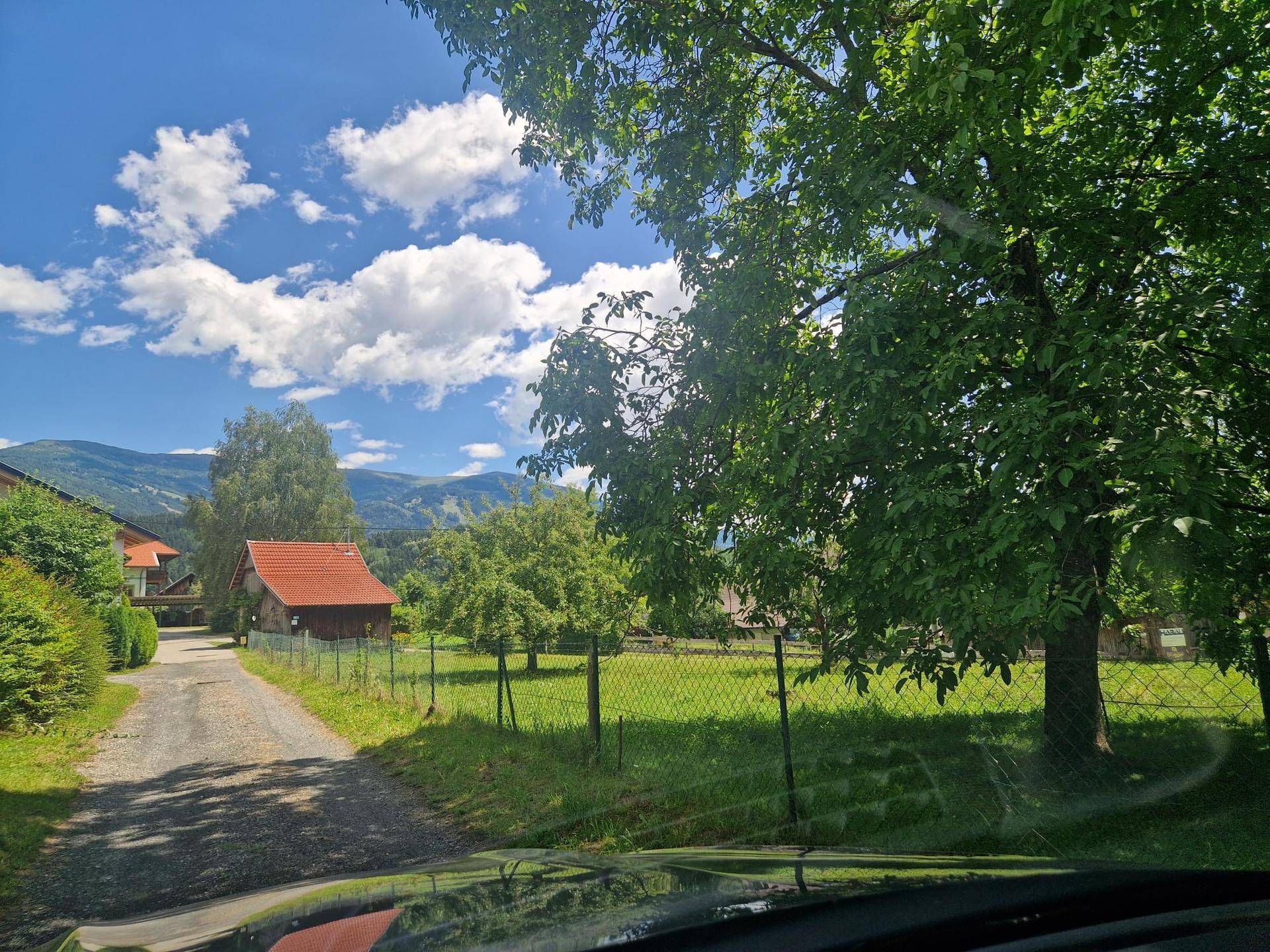 Blick auf eine ländliche Landschaft mit Feldweg, grünen Wiesen, Bäumen und Bergen in der Ferne.