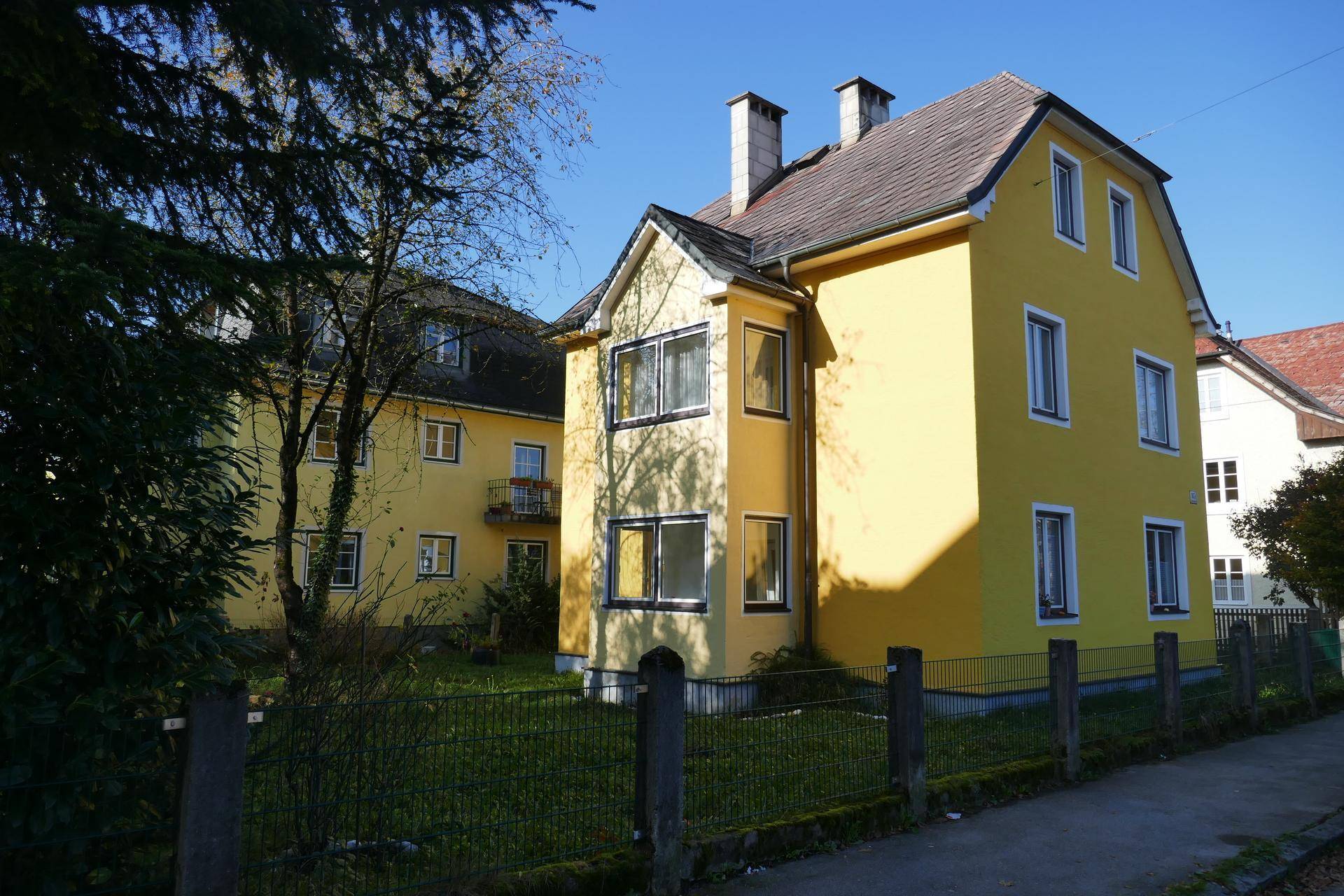 Gelbes Mehrfamilienhaus mit Satteldach und grünem Vorgarten an einem sonnigen Tag.