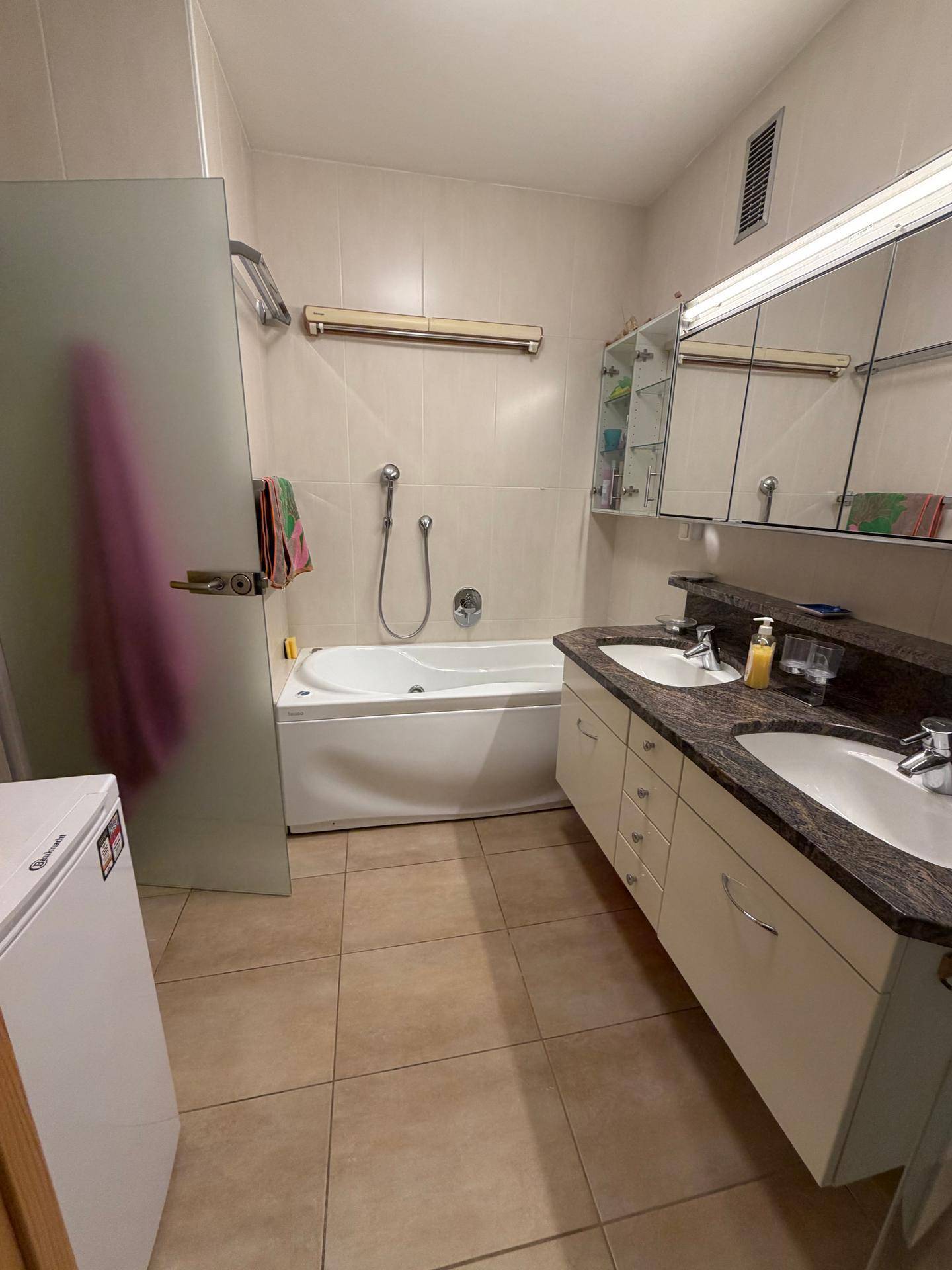 Helles Badezimmer mit Doppelwaschbecken, Badewanne und beigen Fliesen.