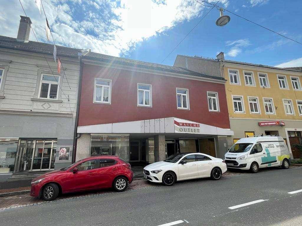 Gesamtansicht der Geschäftsfassade mit parkenden Autos und angrenzenden Gebäuden unter bewölktem Himmel.