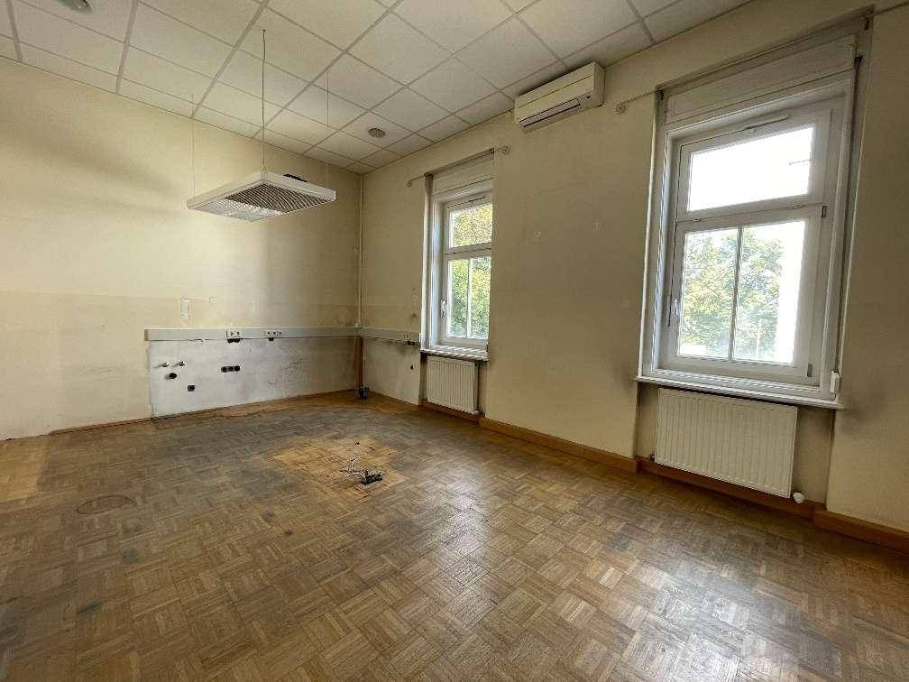 Geräumiger, leerer Raum mit Fischgrätparkett, großen Fenstern und Klimaanlage, bereit zur Renovierung.