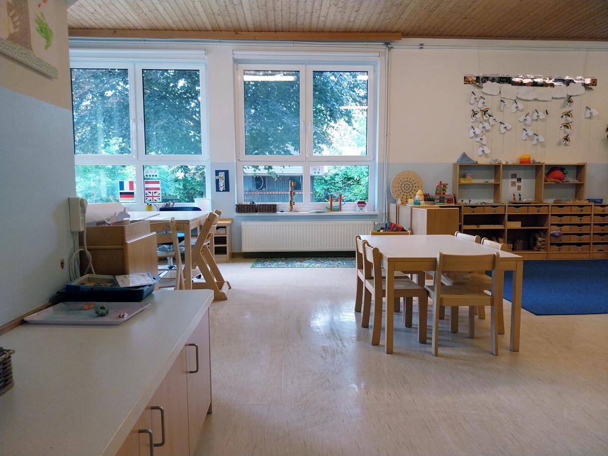 Klassenraum mit Holztischen, Stühlen, Regalen und großen Fenstern mit Blick ins Grüne.