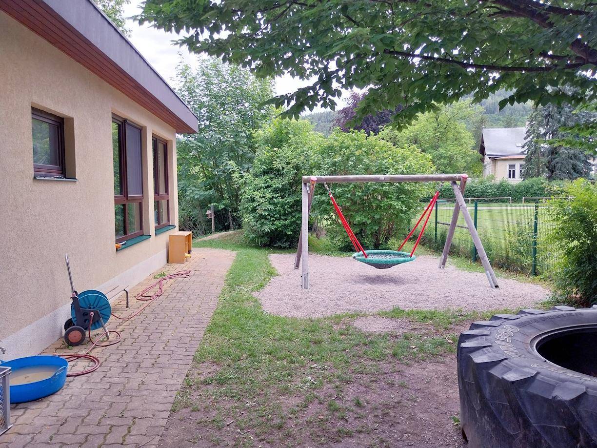 Spielplatzbereich mit Schaukel, Sandfläche und gepflastertem Weg neben dem Gebäude.