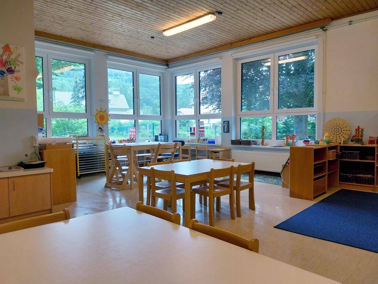 Heller Klassenraum mit Holztischen, Stühlen, großen Fenstern und einer Holzdecke.