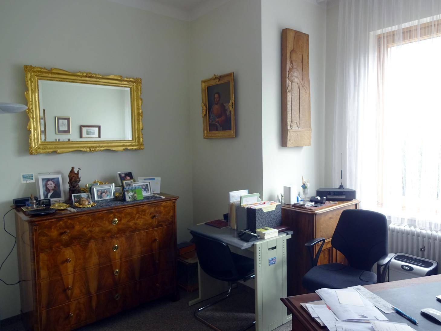 Helles Büro mit klassischer Holzkommode, großem Spiegel und Arbeitsplatz am Fenster.