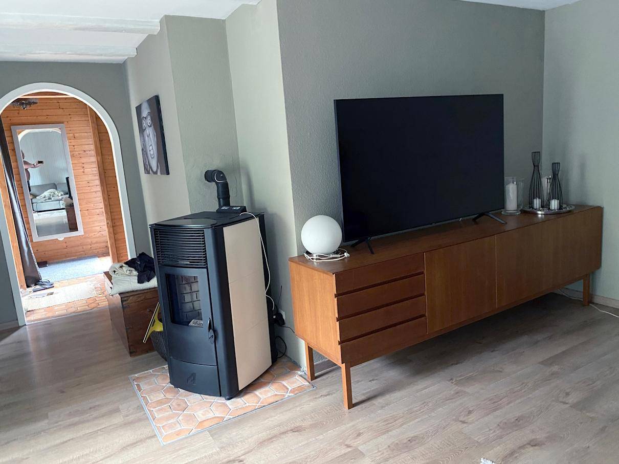 Wohnbereich mit Fernseher, modernem Pelletofen und Holzboden, der zu einem weiteren Raum führt.