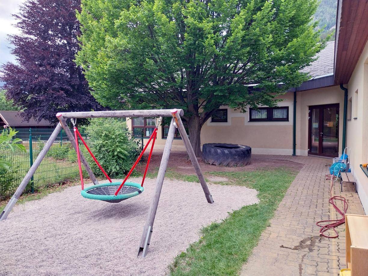 Spielplatzbereich mit Schaukel, Sandfläche und gepflastertem Weg neben dem Gebäude.