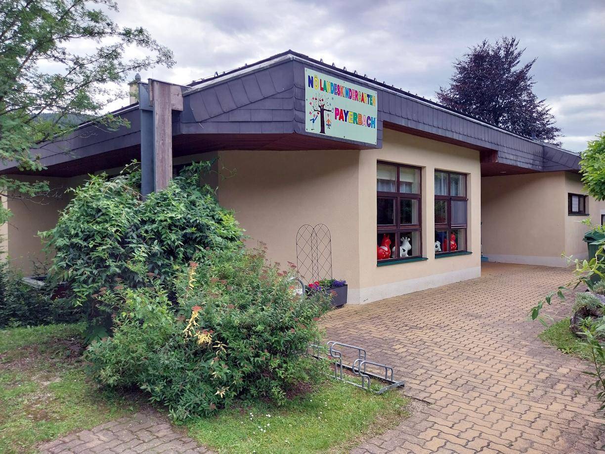 Fassade des Gebäudes mit großen Fenstern, davor gepflasterter Bereich und grüne Büsche.