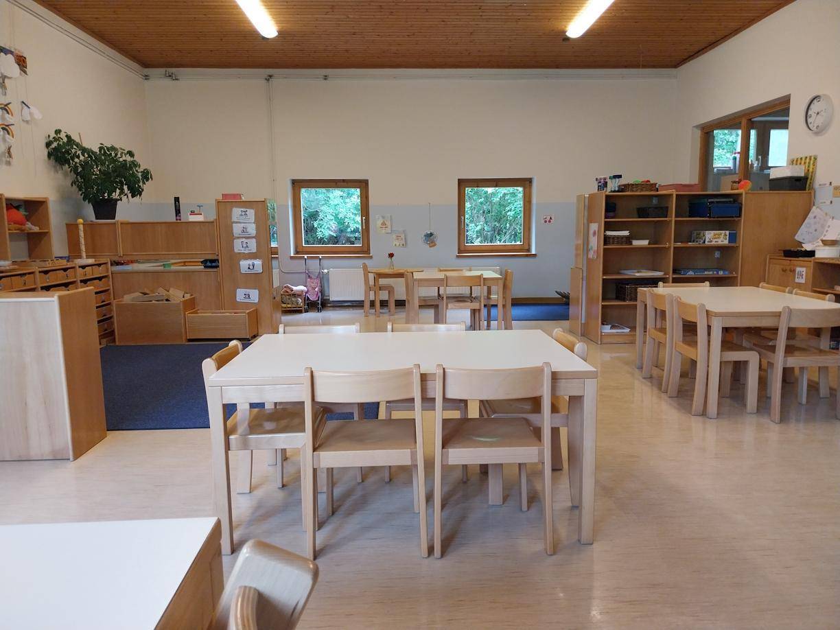 Heller Klassenraum mit Holztischen, Stühlen, Regalen und kleineren Fenstern.