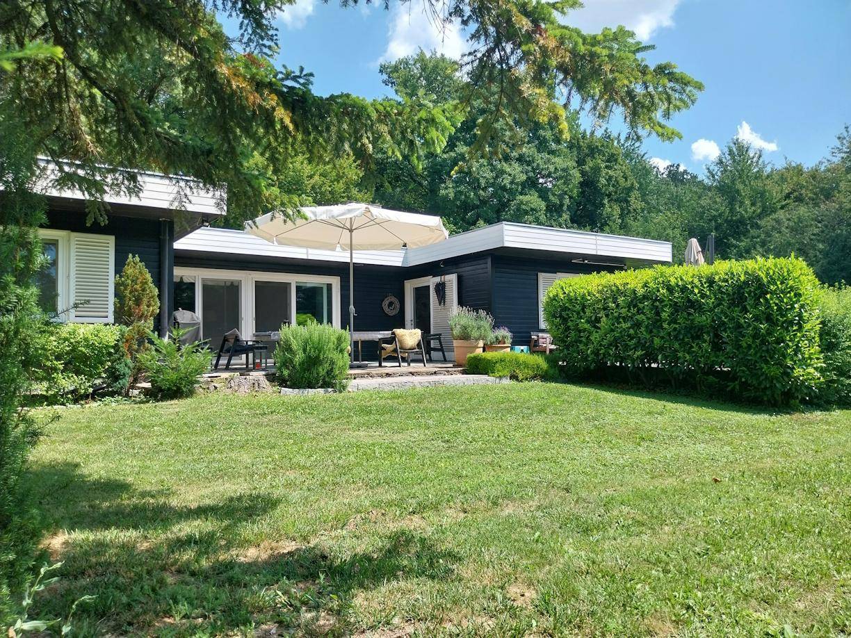 Seitlicher Blick auf das moderne Haus mit großer Terrasse, Gartenmöbeln und umgebender grüner Natur.