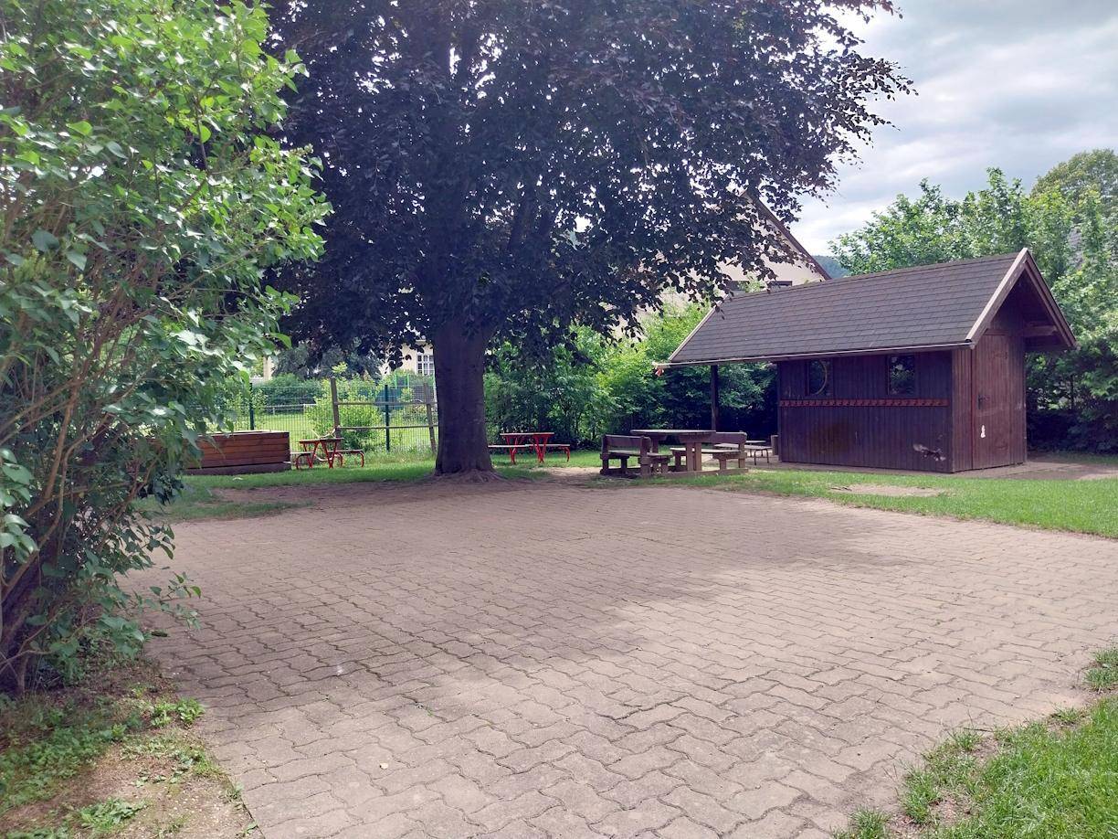 Außenbereich mit gepflasterter Fläche, großem Baum und einem Holzschuppen mit Picknicktischen.