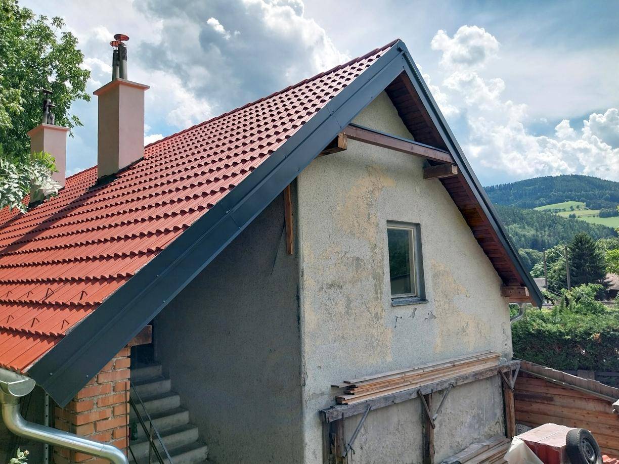 Seitenansicht des Hauses mit rotem Ziegeldach und Schornstein vor bewaldeter Hügellandschaft.