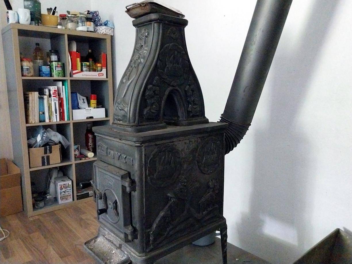 Historischer Gusseisenofen mit Verzierungen und einem Regal mit Haushaltsartikeln im Hintergrund.