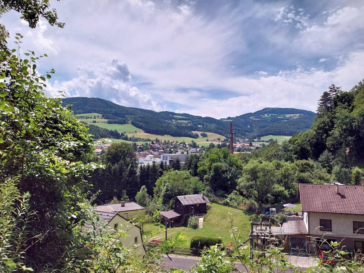Weitläufiger Blick über eine ländliche Siedlung mit bewaldeten Hügeln und einem Industrieschornstein in der Ferne.