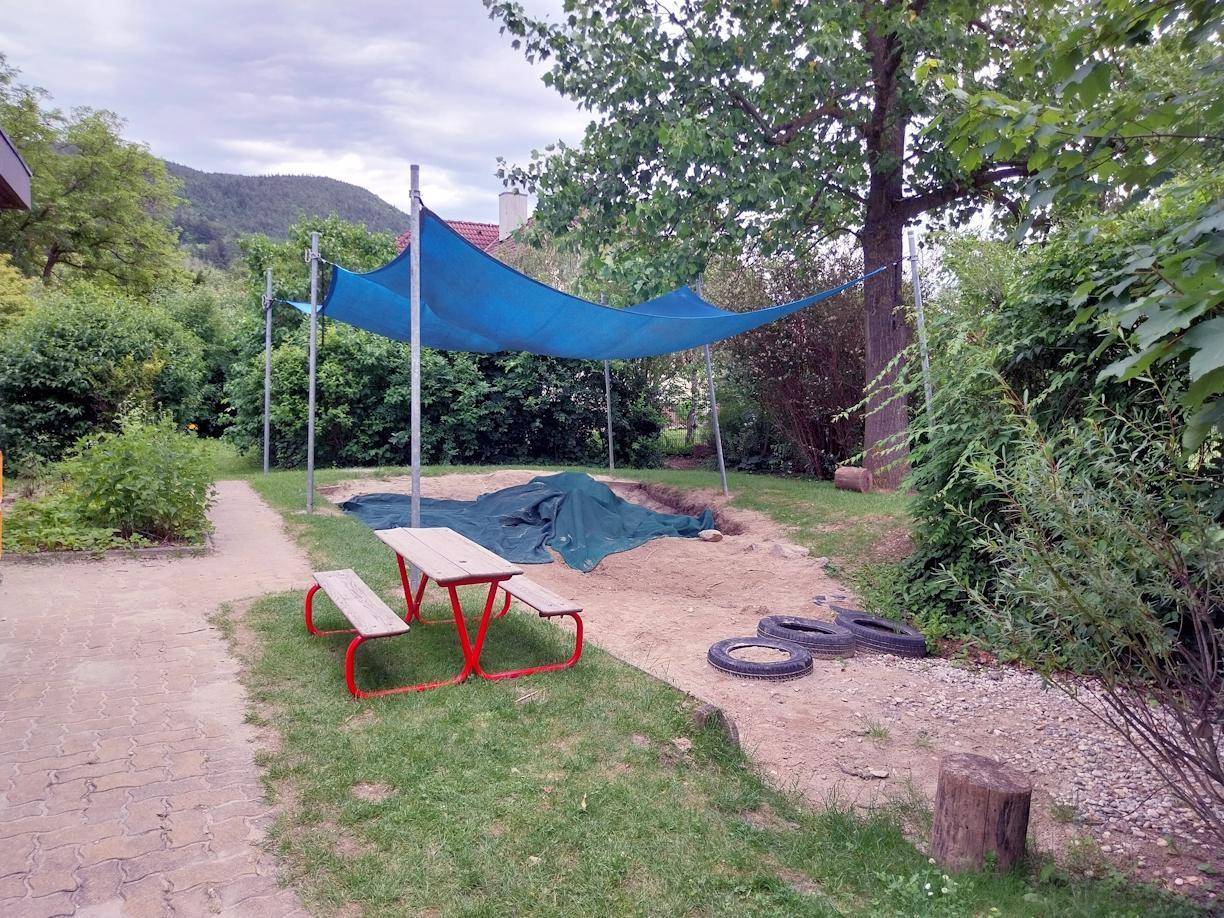Spielplatzbereich mit Sandkasten unter blauem Sonnensegel und kleinem Picknicktisch.