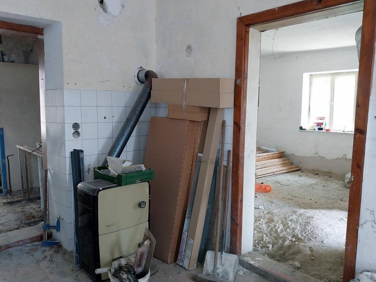Renovierungsbedürftiger Raum mit einem Ofen, gefliesten Wänden und einem Durchgang zu einem weiteren Zimmer.