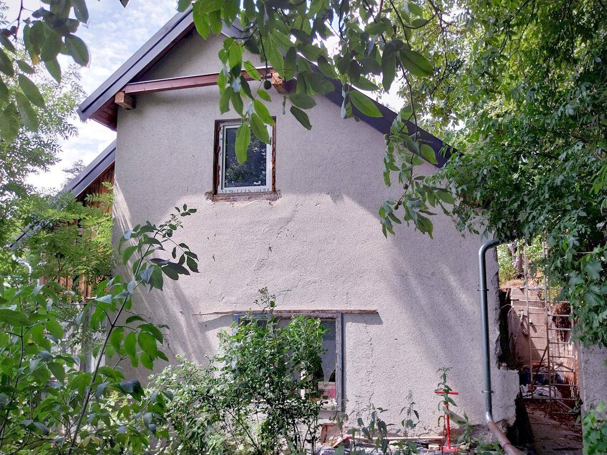 Seitenansicht des Hauses mit einfacher Fassade, teilweise von dichtem Grün und Bäumen verdeckt.