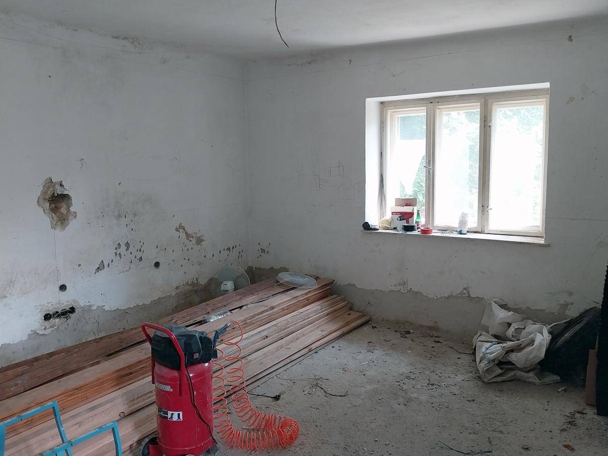 Renovierungsbedürftiger Raum mit abblätternder Farbe, Holzdielen und einem Fenster, bereit für die Sanierung.