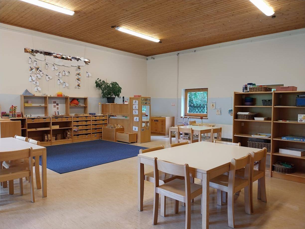 Geräumiger Klassenraum mit Holzregalen, Tischen, Stühlen und einem blauen Teppichbereich.