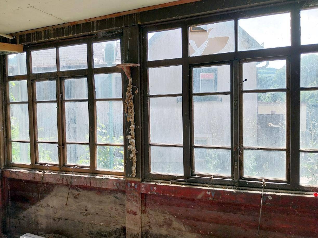 Reihe alter Holzfenster mit Sprossen und teilweise beschädigtem Glas in einem renovierungsbedürftigen Raum.