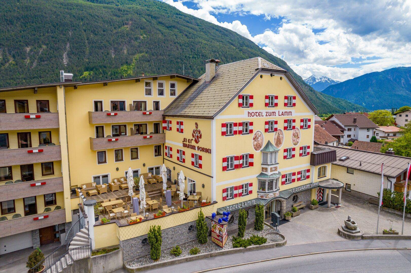 Ansicht des Hotels mit Außenterrasse und traditioneller Architektur, umgeben von grünen Bergen und blauem Himmel.