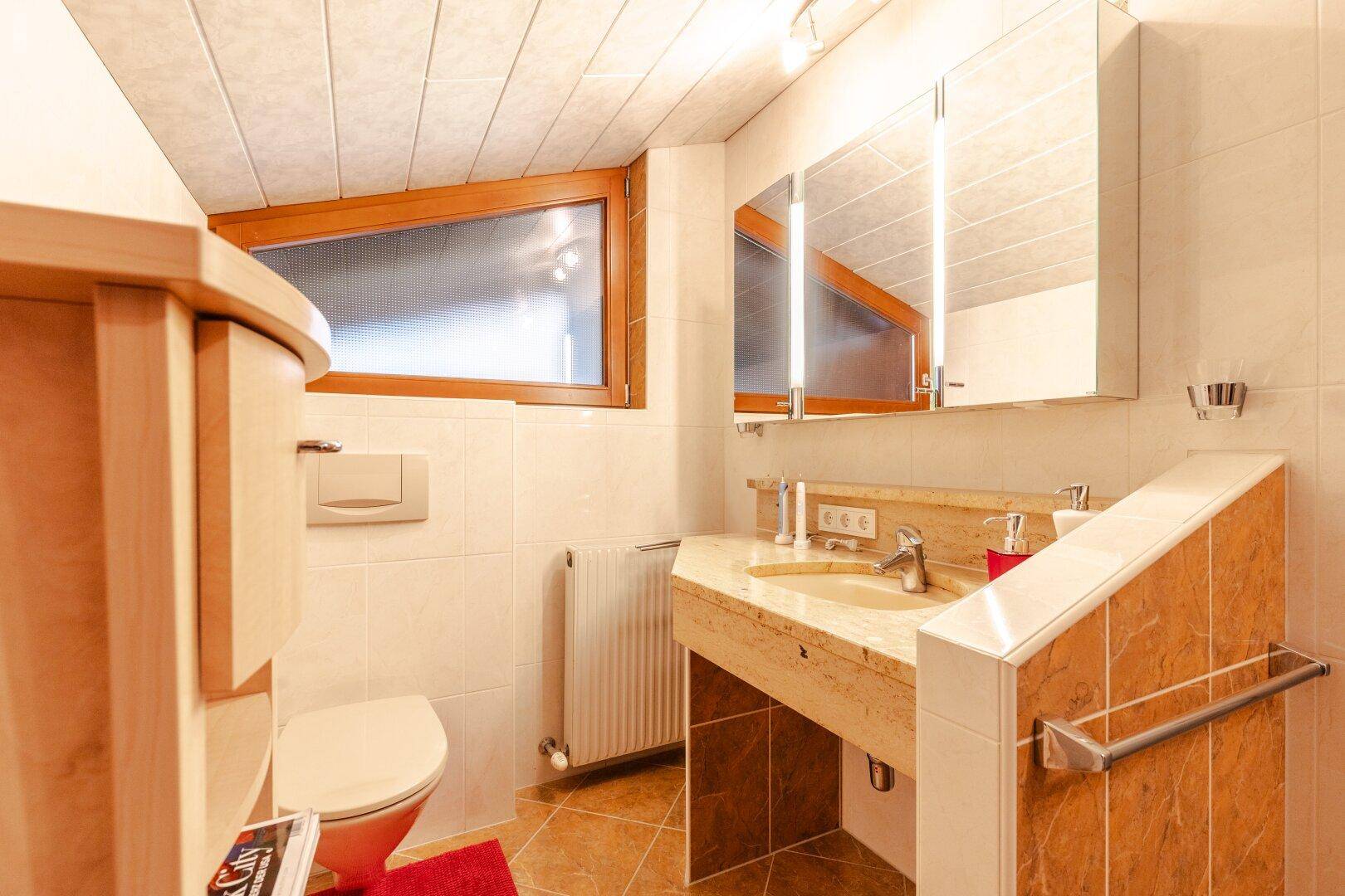 Badezimmer mit Waschtisch, WC, Spiegelschrank und Fenster.