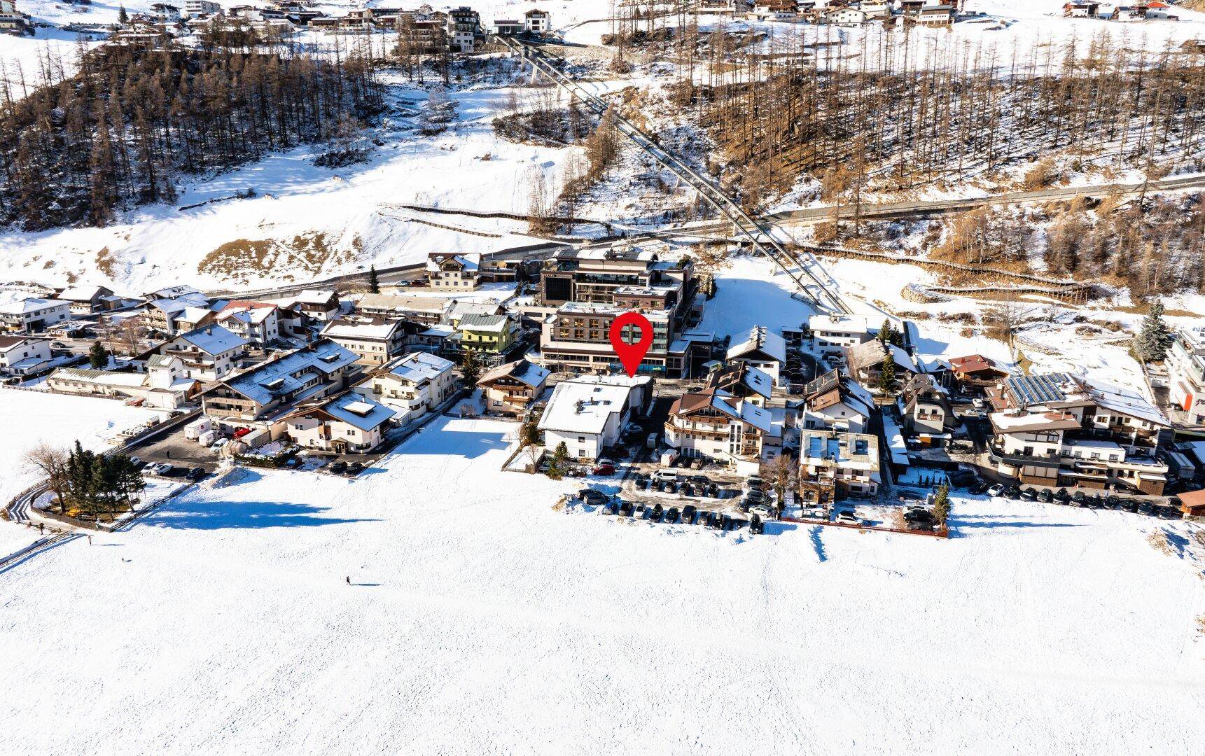Luftaufnahme eines Stadtteils mit weitem, schneebedecktem Feld im Vordergrund und Gebäuden, eingebettet in eine Winterlandschaft.