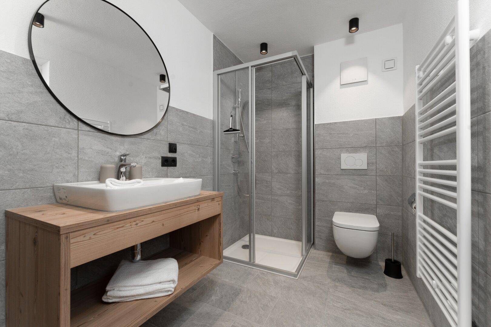 Modernes Badezimmer mit begehbarer Dusche, Holztisch und beheiztem Handtuchhalter, in hellen Grautönen gehalten.