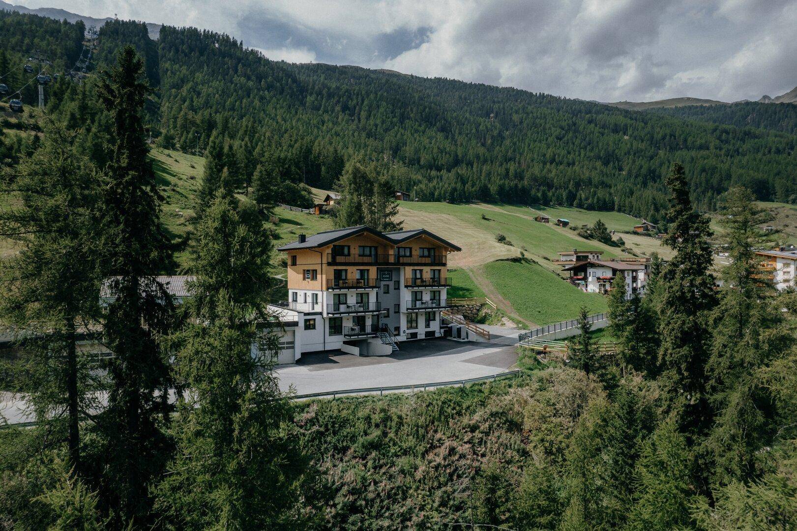 Das Apartmentgebäude ist eingebettet in die grüne Berglandschaft, umgeben von hohen Bäumen und Wiesen.