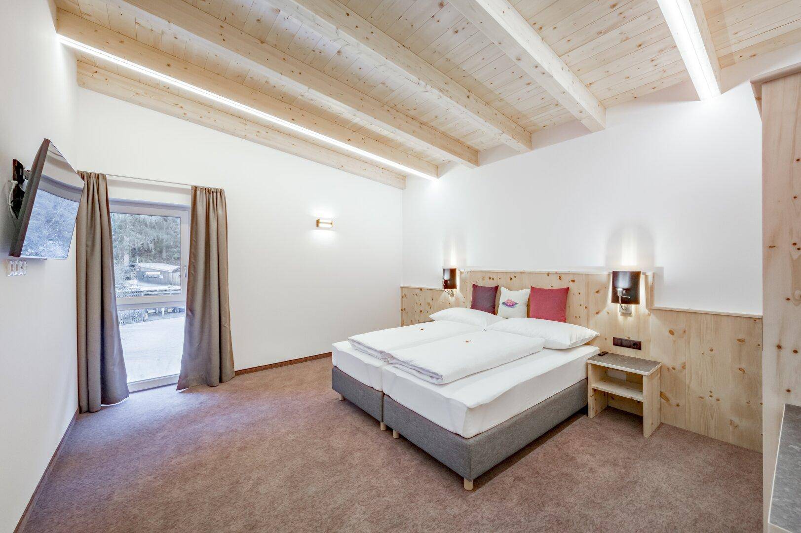 Helles Schlafzimmer mit Holzbalkendecke, Doppelbett und Zugang zu einem Außenbereich durch eine große Tür.