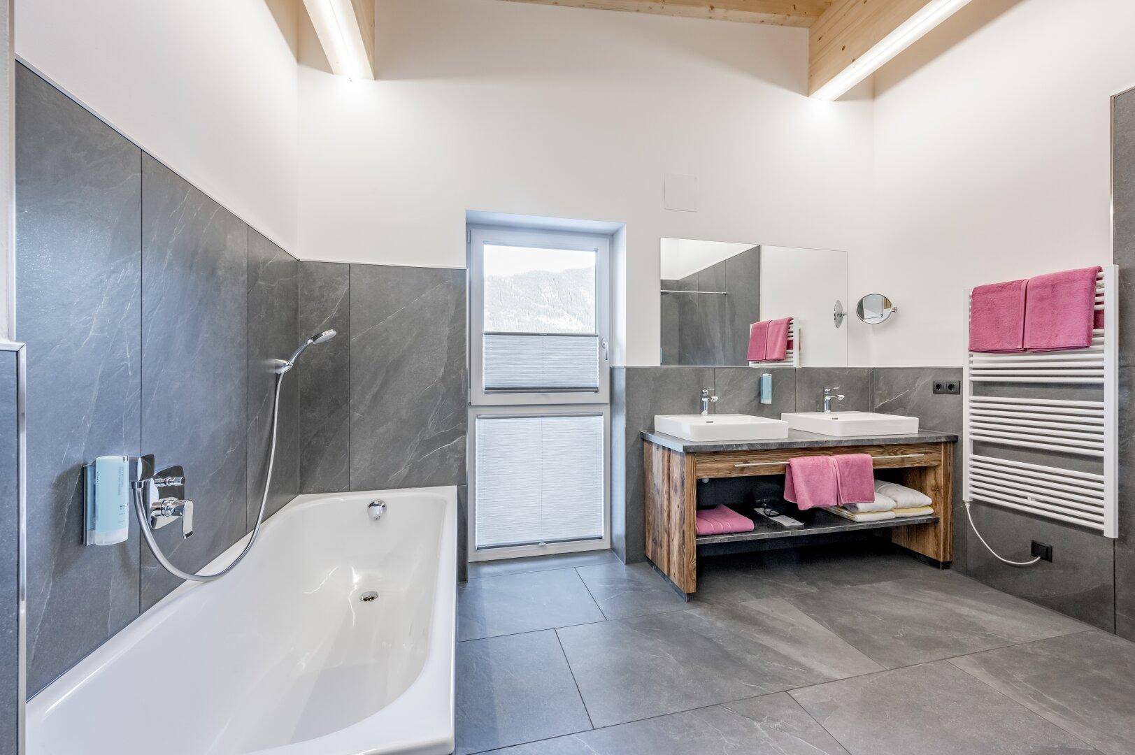 Geräumiges Badezimmer mit Badewanne, Doppelwaschbecken, großem Spiegel und einem Fenster für Tageslicht.