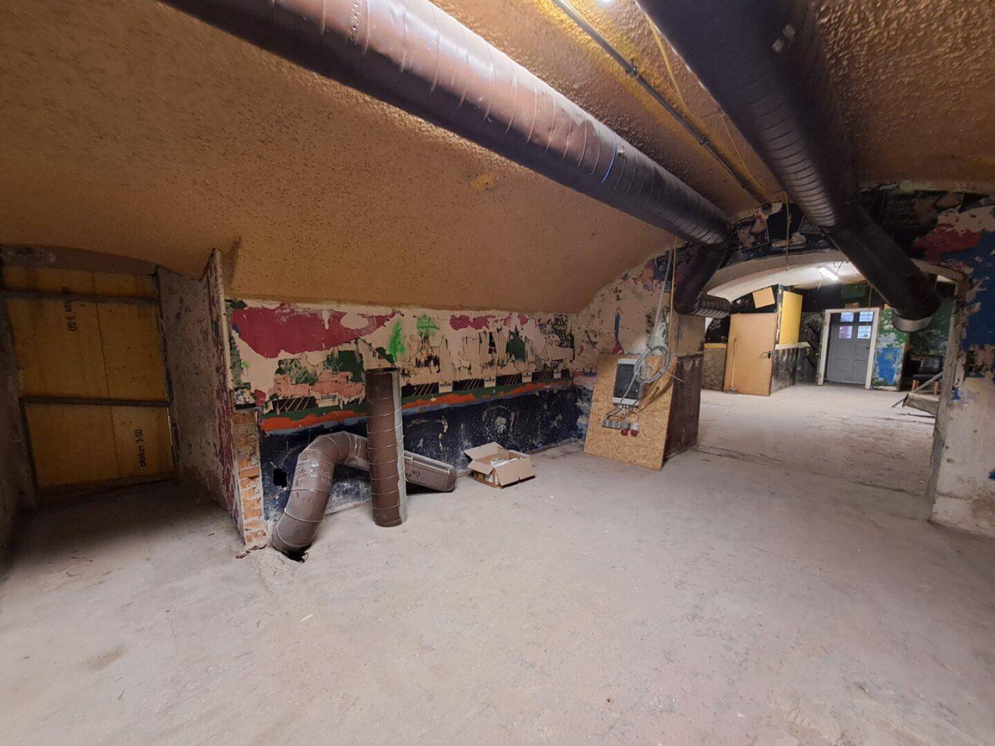 Großer Kellerraum mit gewölbter Decke, sichtbaren Leitungen und Spuren von Verfall.