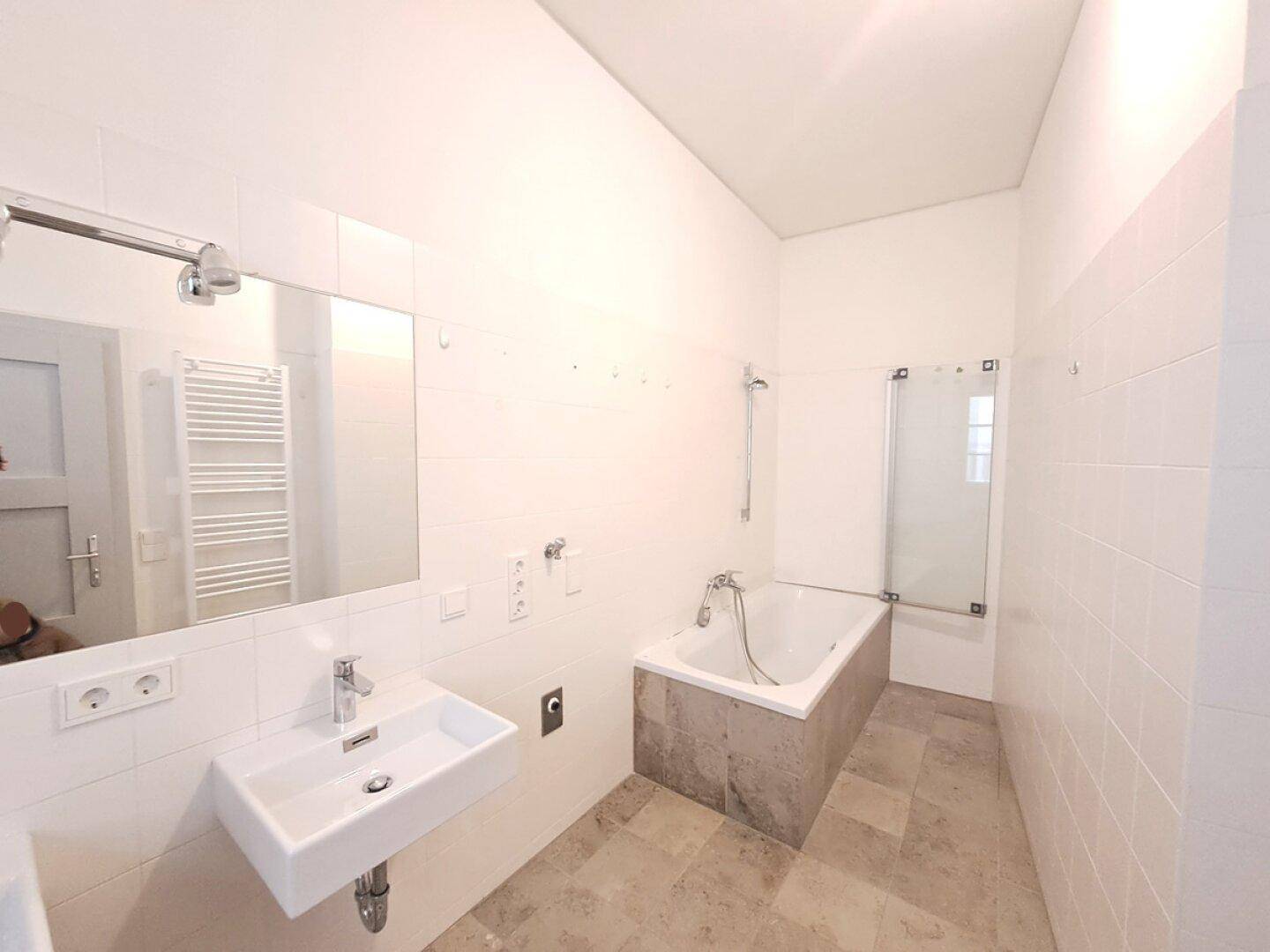 Helles Badezimmer mit weißen Fliesen, Badewanne, modernem Waschbecken und beheiztem Handtuchhalter.