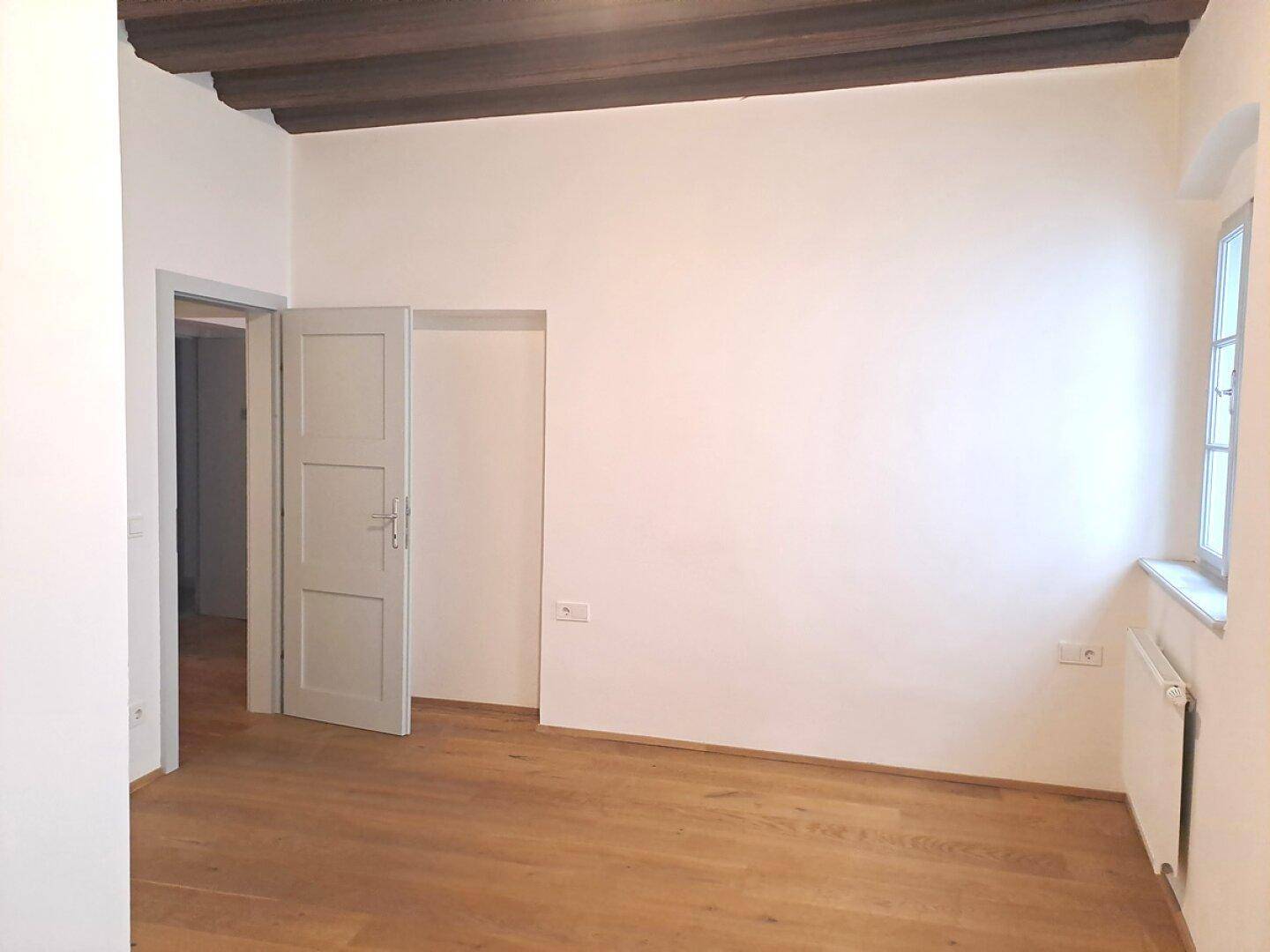 Heller Raum mit Holzbalkendecke, Dielenboden und einem Fenster, das Tageslicht spendet.