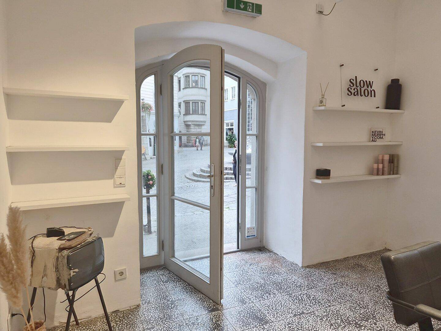 Eingangsbereich eines Salons mit offener Glastür, die den Blick auf eine historische Gasse freigibt.