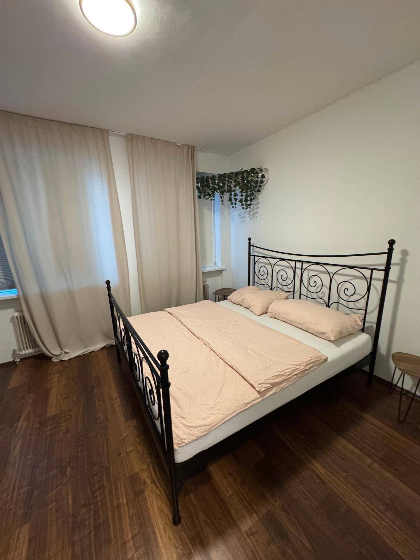 Schlafzimmer mit schmiedeeisernem Bettgestell, hellen Vorhängen und dunklem Holzfußboden.