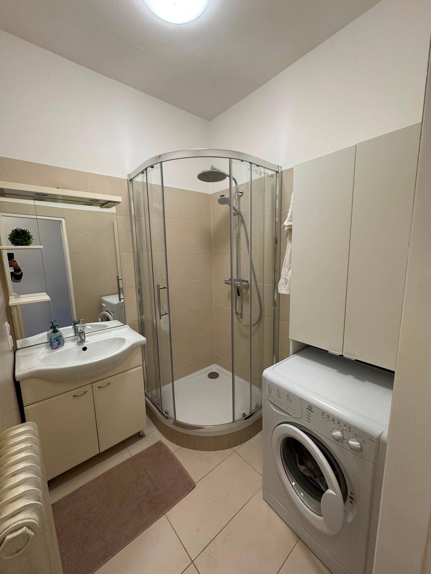 Badezimmer mit Eckdusche, Waschbecken und Waschmaschine, hell gefliest.