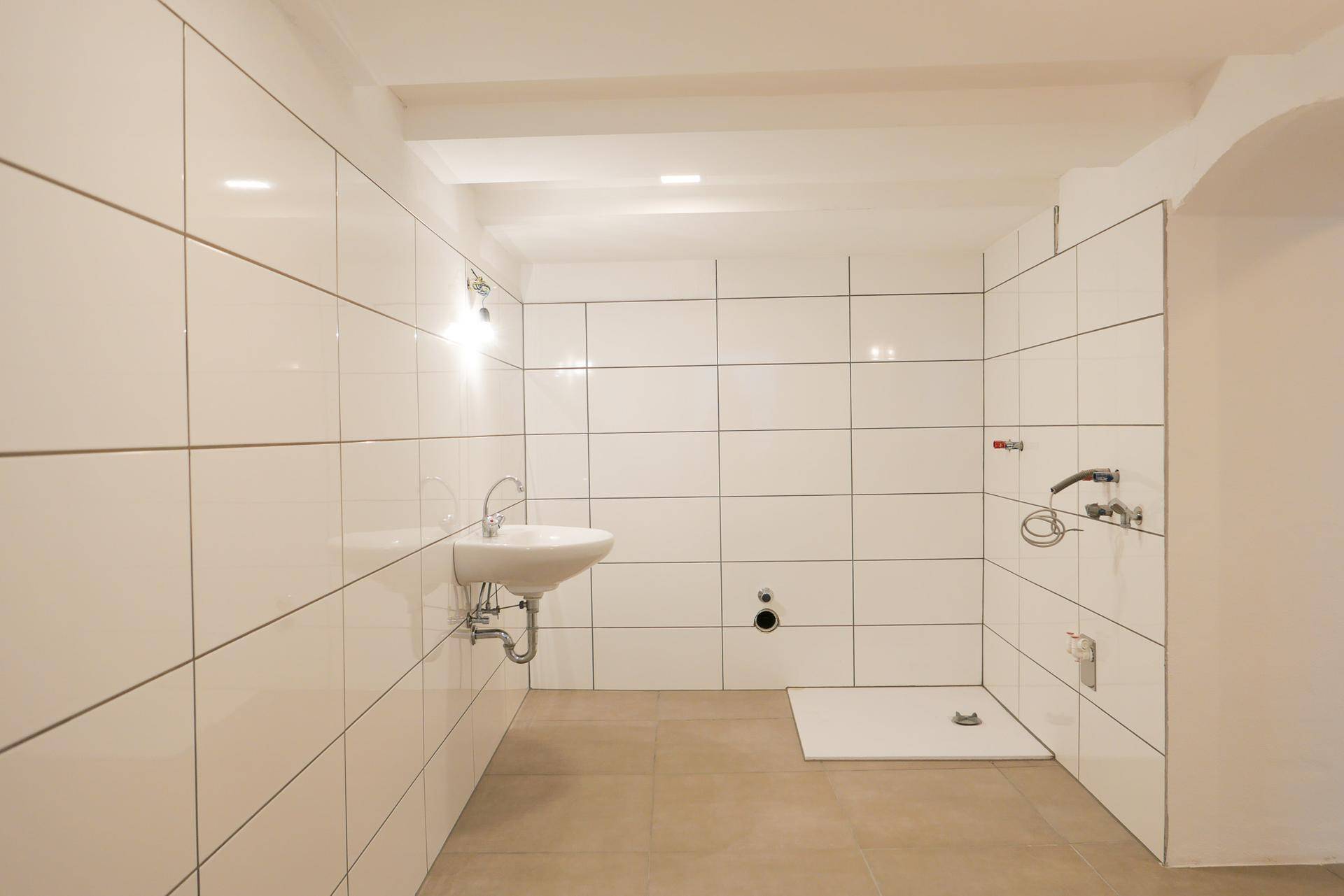 Helles Badezimmer mit weißen Wandfliesen, einem Waschbecken und einem Duschbereich mit Bodenablauf.