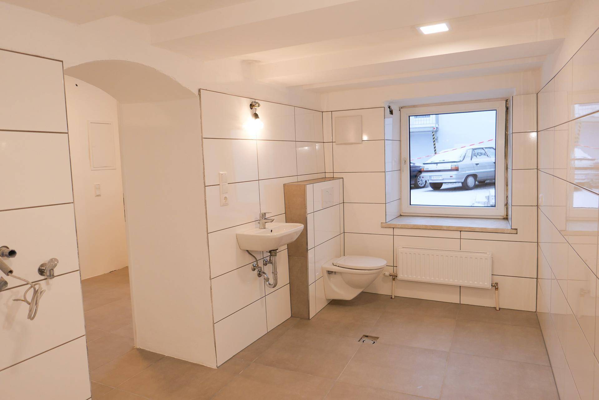 Modernes Badezimmer mit weißen Fliesen, wandhängender Toilette, Waschbecken und einem Fenster mit Straßenblick.