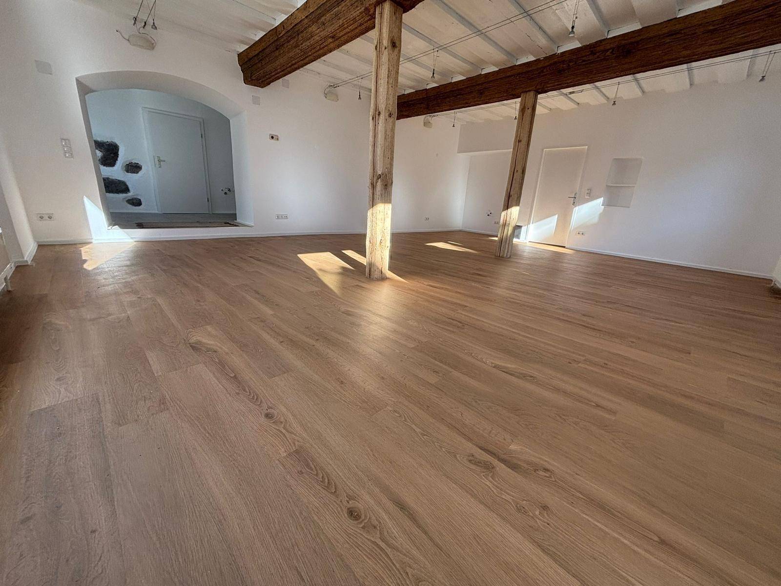 Großer, heller Raum mit Holzboden, freiliegenden Holzbalken und einem offenen Durchgang zu einem weiteren Bereich.