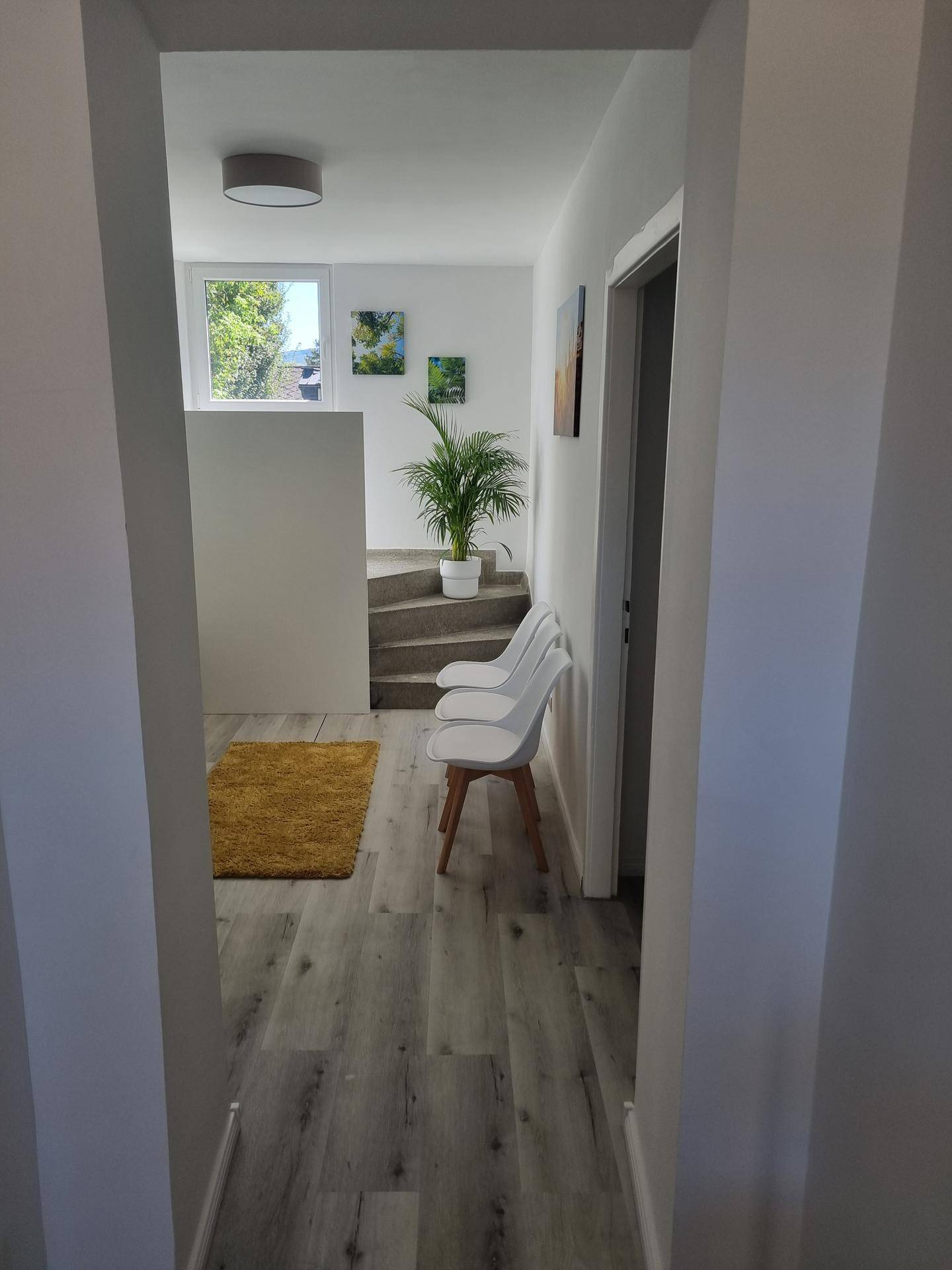 Heller Wartebereich mit weißen Stühlen, Laminatboden und einer Treppe, die zu einem Fenster führt.