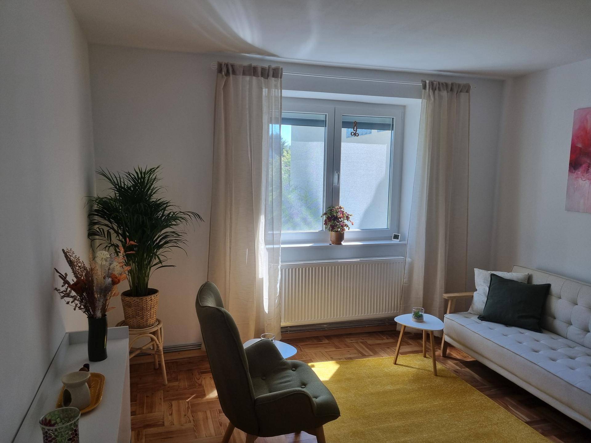 Gemütlicher Raum mit Fischgrätparkett, einem beigen Sofa, einem Sessel und einem großen Fenster mit Heizkörper.