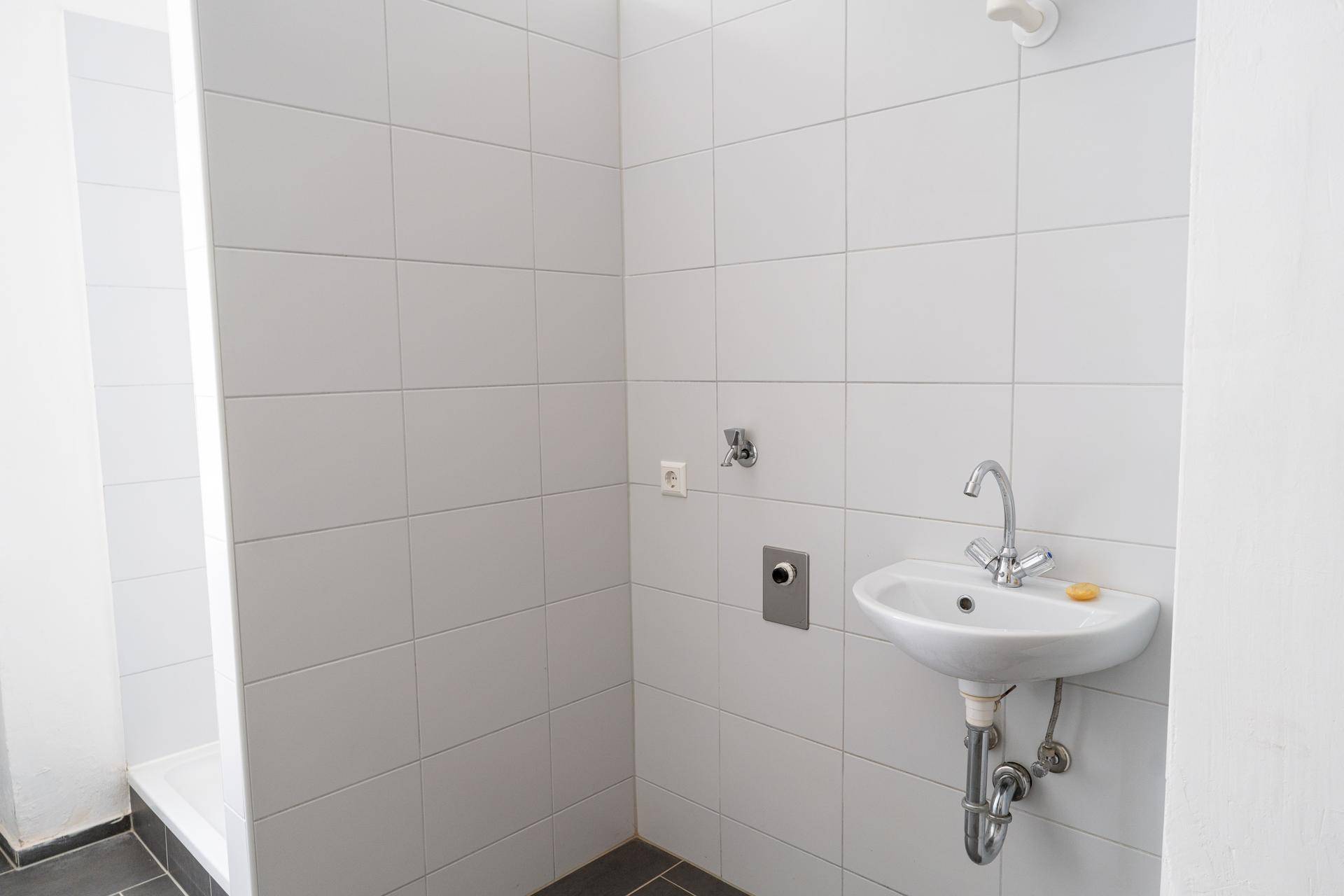 Helles Badezimmer mit weißen Fliesen, kleinem Waschbecken und einer Duschgelegenheit.