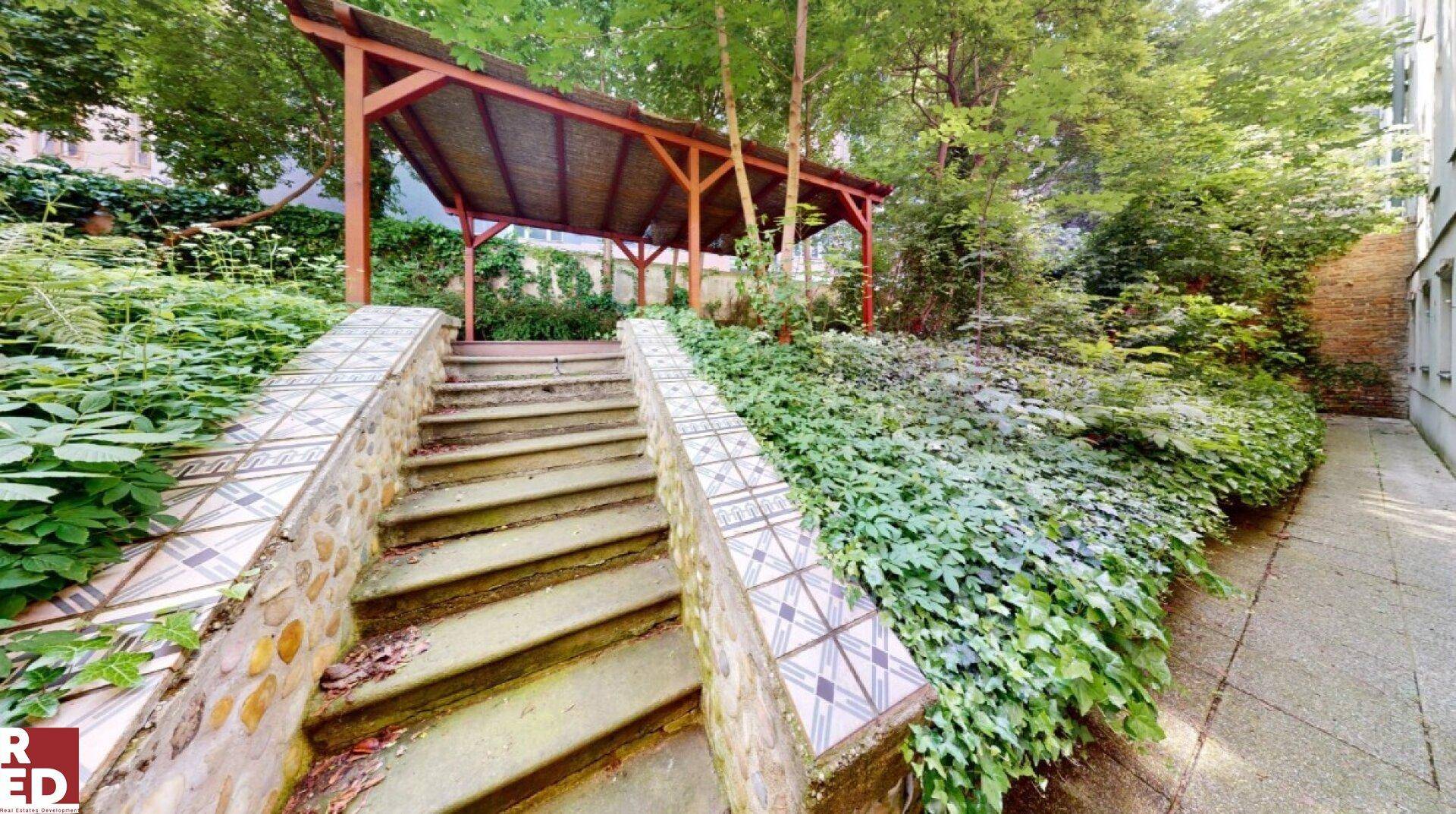 Grüner Gartenbereich mit Steintreppe, üppiger Bepflanzung und einem überdachten Sitzbereich im Hintergrund.