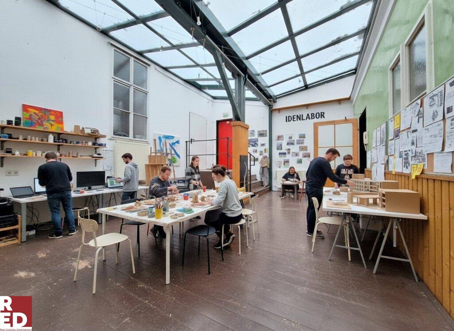 Kreativer Arbeitsbereich mit Glasdach, Holzboden und mehreren Personen, die an Tischen arbeiten.