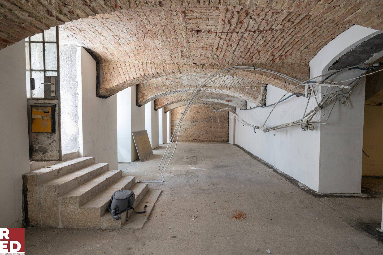 Geräumiger Kellerraum mit freiliegenden Ziegelgewölben, Betonboden und einer Steintreppe.