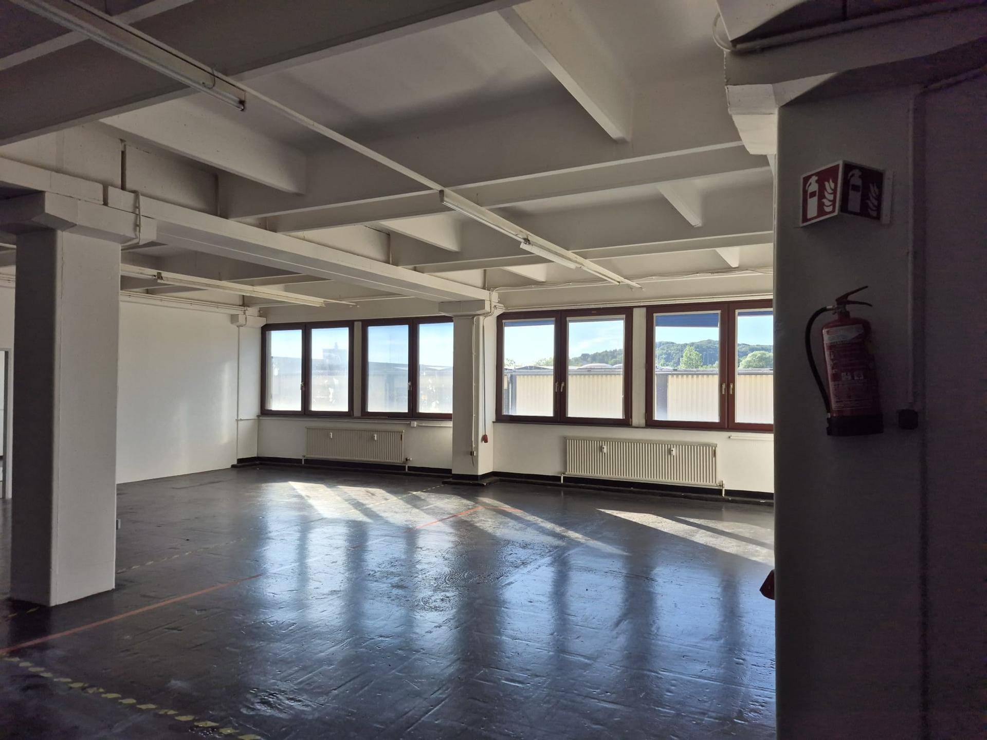 Großer Lagerraum mit dunklem Betonboden, sichtbaren Deckenbalken und vielen Fenstern.