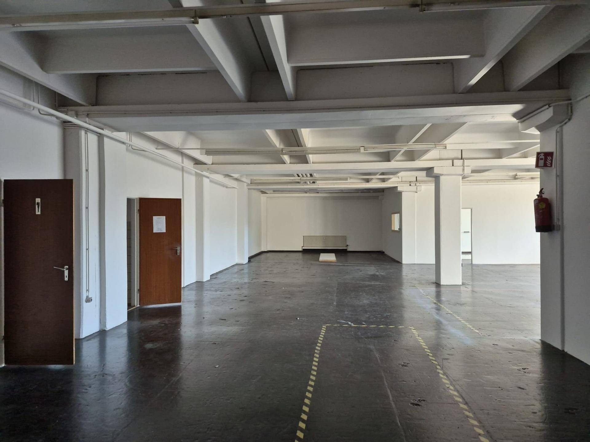 Großer Lagerraum mit dunklem Betonboden, hohen Decken und zwei braunen Türen.