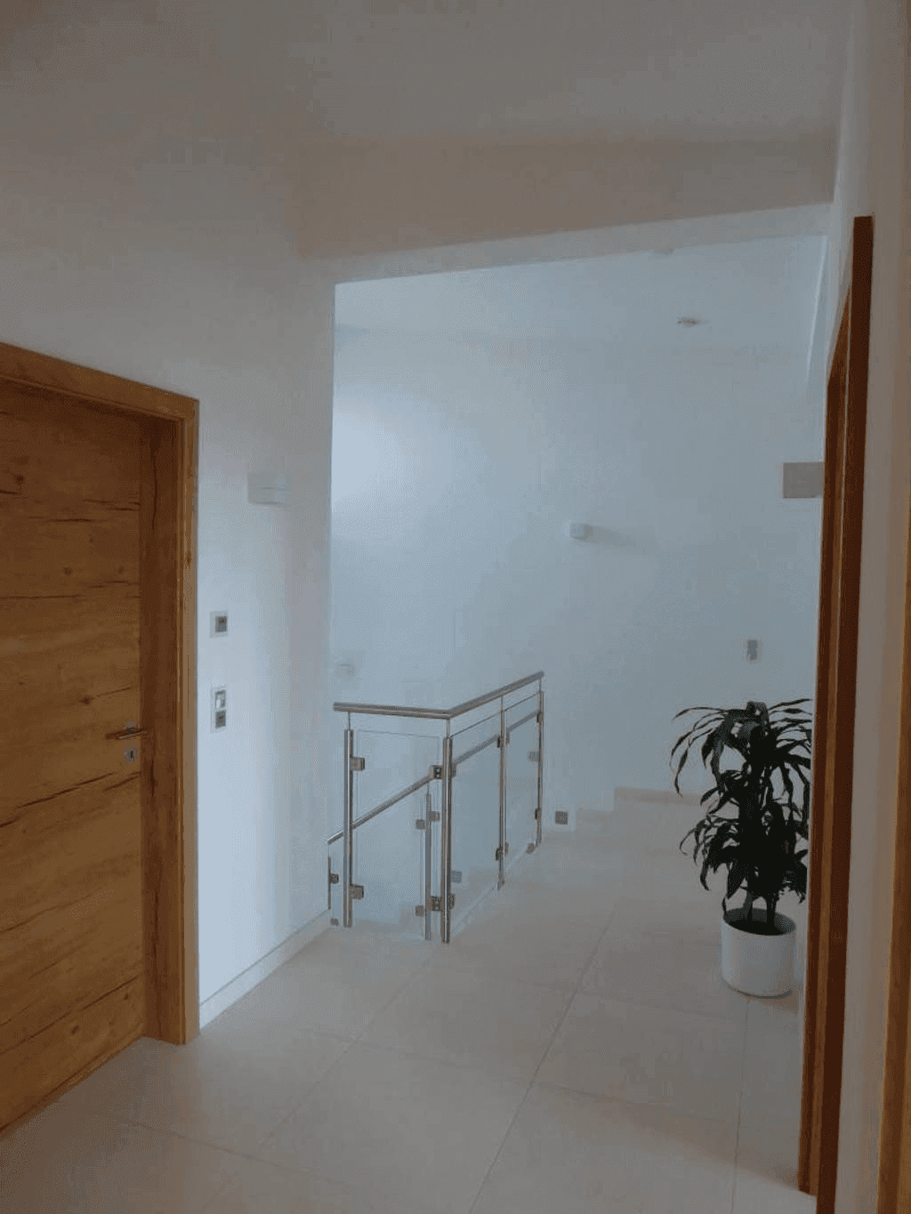 Heller Flur mit Fliesenboden, Holztreppe und Glasgeländer, sowie einer Zimmerpflanze.