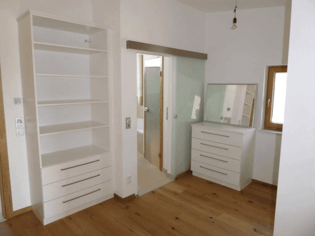 Helles Schlafzimmer mit Holzboden, weißem Regal und Kommode, sowie Schiebetür zum Bad.